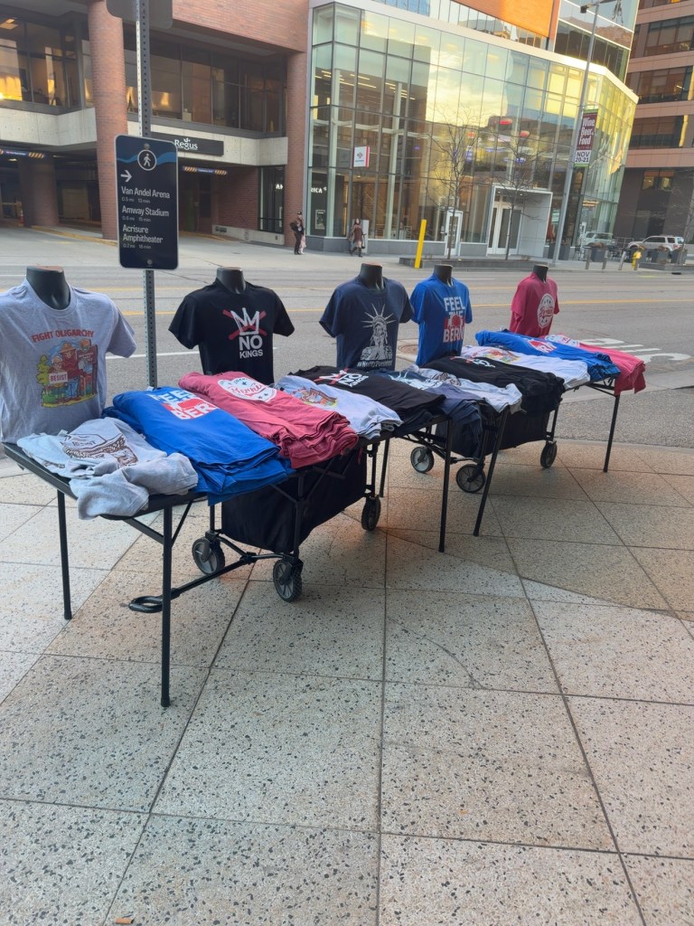Tables of graphic T-shirts displayed on a downtown Grand Rapids sidewalk, including No Kings and Feel the Bern designs.