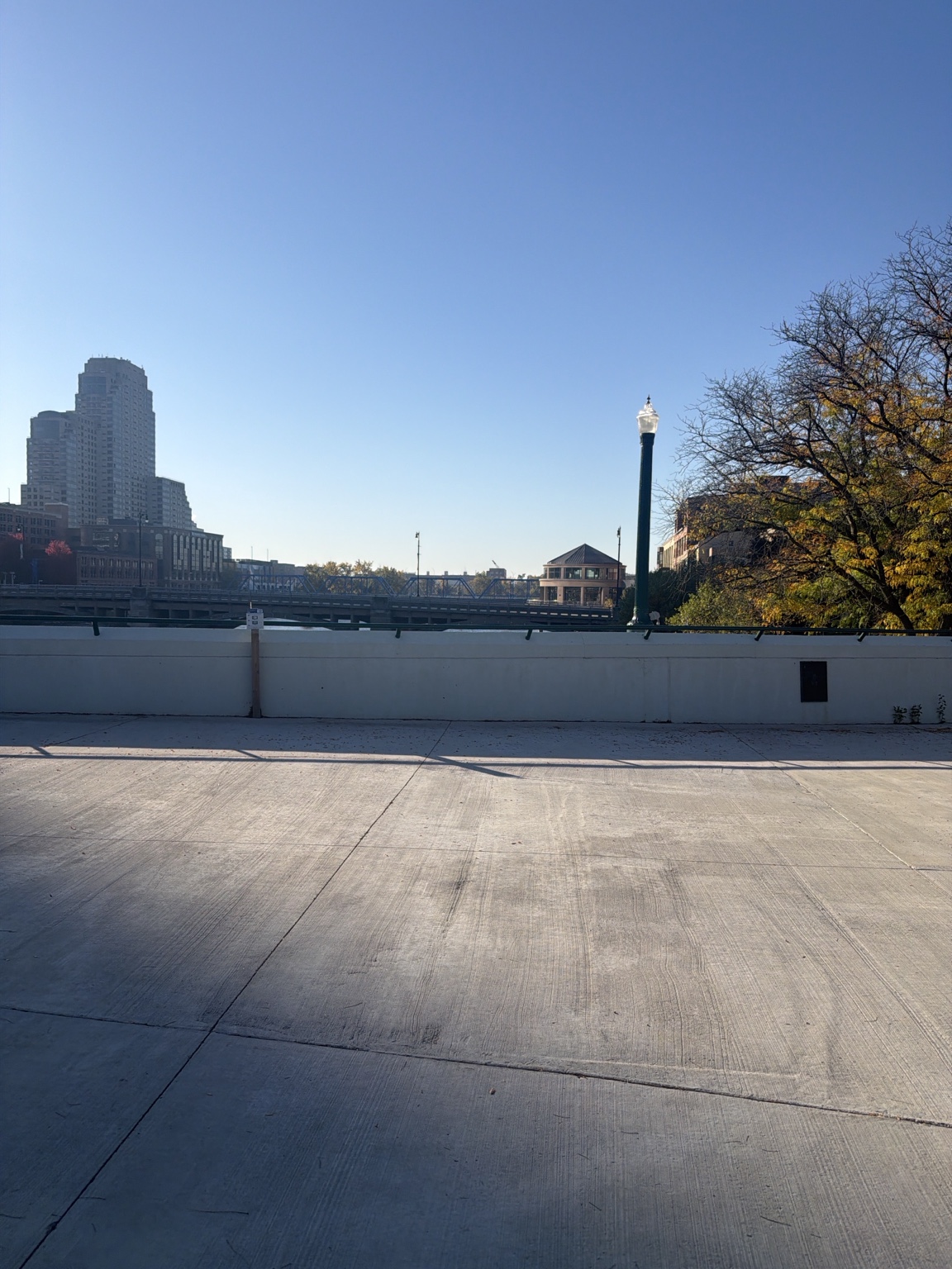 A paved riverside area with trees and city buildings beyond.